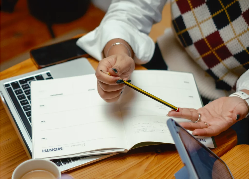 Melin's hands holding a pencil while planning in an open notebook. The notebook is laid on top of a laptop, and a tablet and a cup of coffee are visible on the wooden desk. The scene suggests a workspace or planning session.