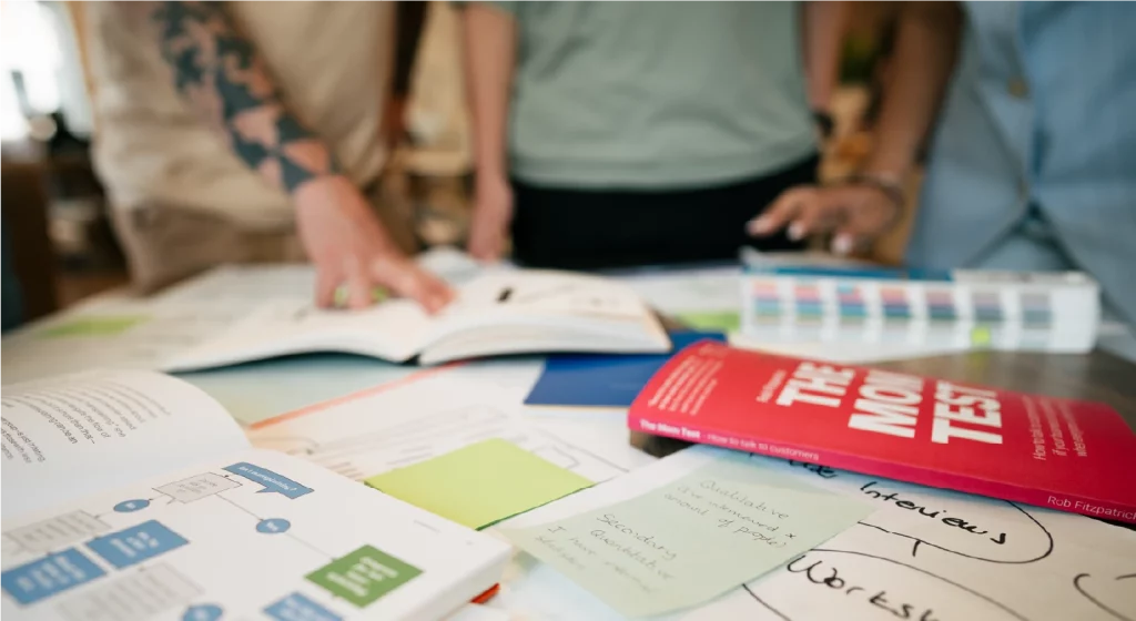 A close-up photograph of a workshop table filled with design strategy materials, including handwritten sticky notes, user flowcharts, and a book titled The Mom Test.