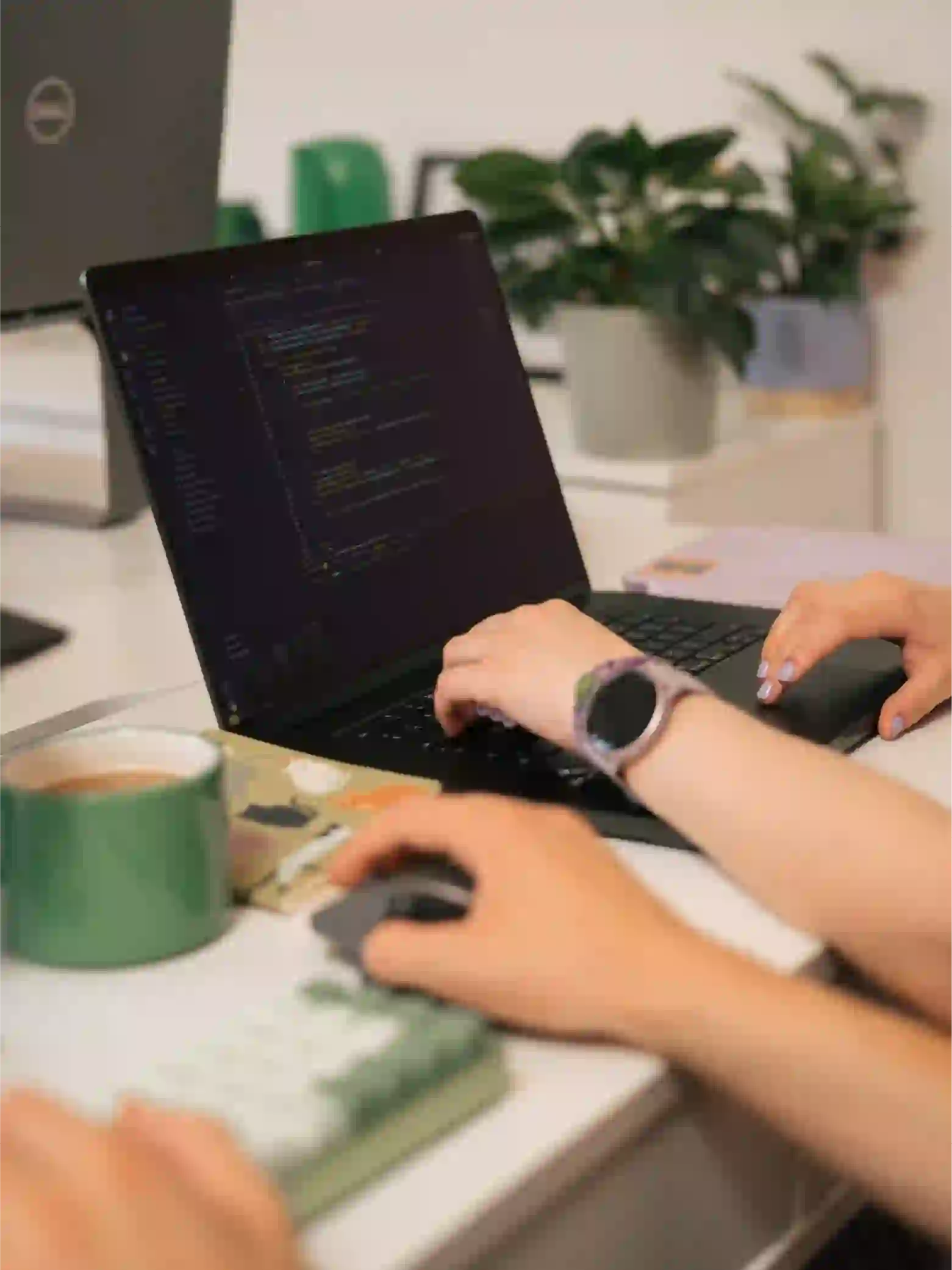 A close-up shot of two developers pair-programming at a white desk, with hands typing code on a dark-mode laptop screen next to a green coffee mug.