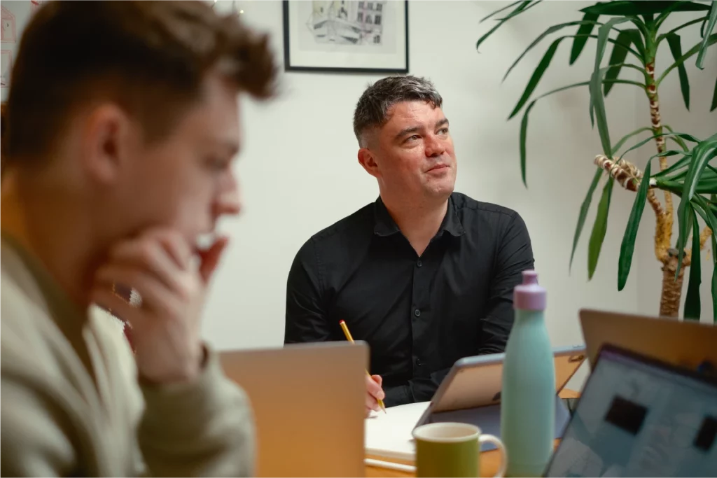 A photograph of a director and a designer during a team meeting, with one holding a pencil and listening thoughtfully at a table with laptops, a tablet, and a coffee mug.