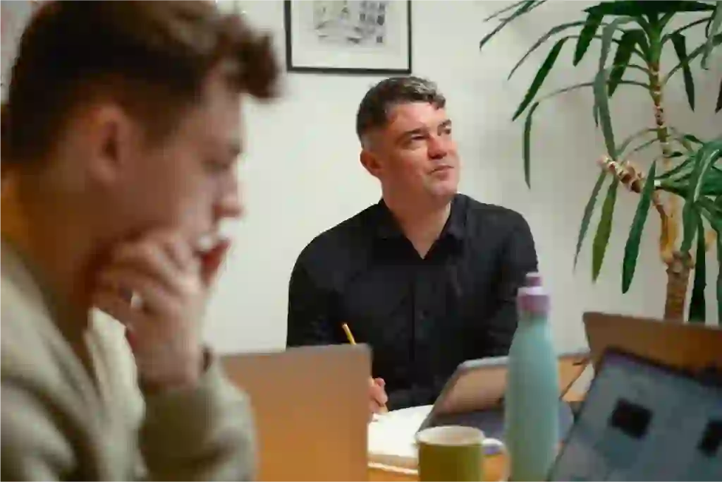 A photograph of a director and a designer during a team meeting, with one holding a pencil and listening thoughtfully at a table with laptops, a tablet, and a coffee mug.